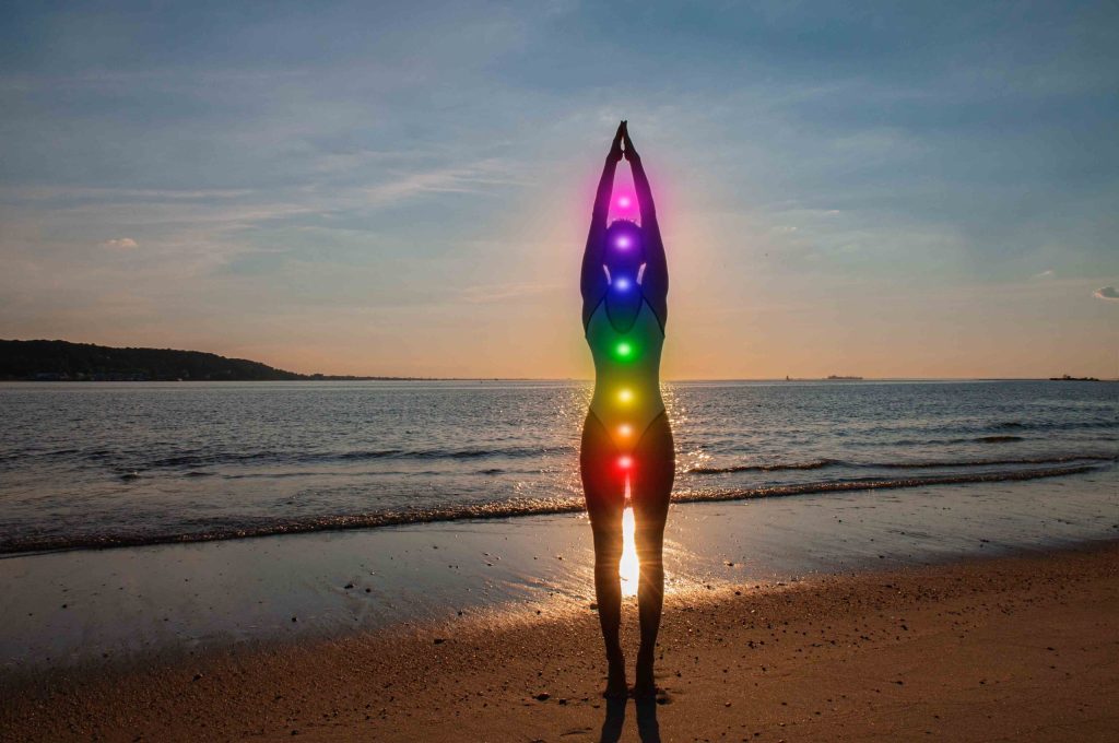 Woman is meditating with glowing seven chakras on the beach. Silhouette of woman is practicing yoga at sunset on sea shore. Kundalini meditation.
